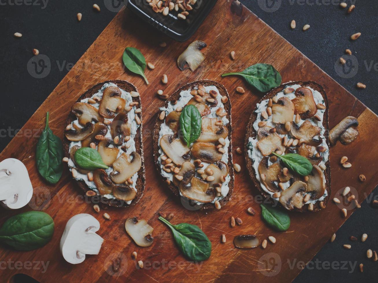 leckere frische Bruschetta mit Pilzen, Spinat, Knoblauch, Frischkäse und Pinienkernen, auf einem Holzbrett, auf dunklem Hintergrund. foto