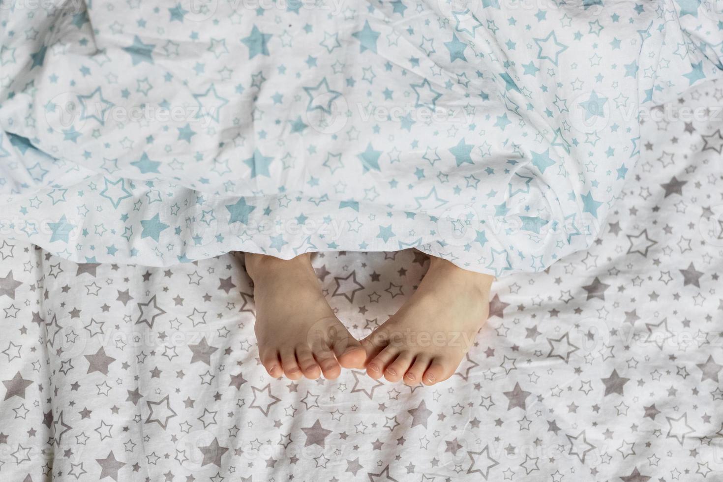 Nahaufnahme der Füße des Kleinkindmädchens auf dem Bett unter der Decke. helle Blau- und Beigetöne. foto