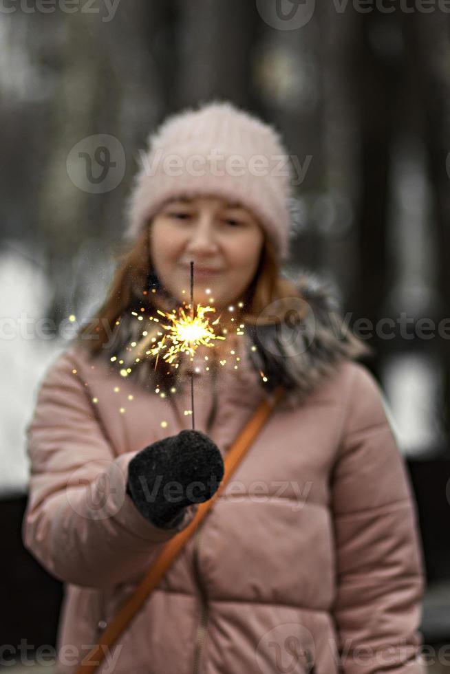 Weihnachtslichter sind helle Spritzer, die in den Händen einer verschwommenen glücklichen Frau im Park brennen. Wunderkerze. Emotionen, Neujahrsstimmung.Winterferien foto