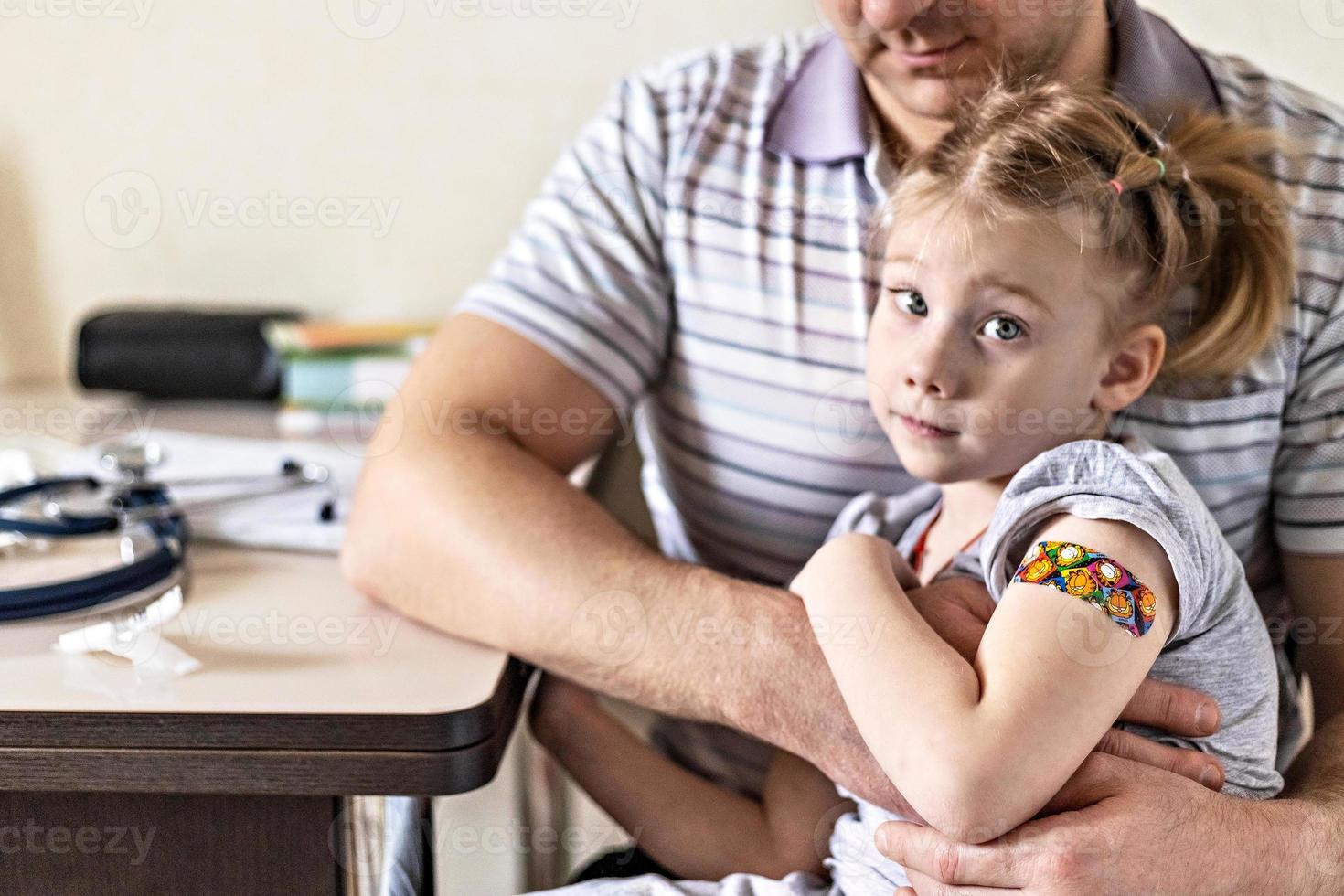 Impfung eines kleinen Mädchens in den Armen ihres Vaters in der Arztpraxis gegen das Coronavirus. lustiges klebepflaster für kinder. Impfung gegen Covid-19, Grippe, Infektionskrankheiten. foto