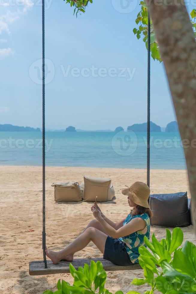 junge Frau, die auf einer Schaukel in der Nähe des Strandes sitzt und während der Sommerferien mit dem Smartphone Fotos macht