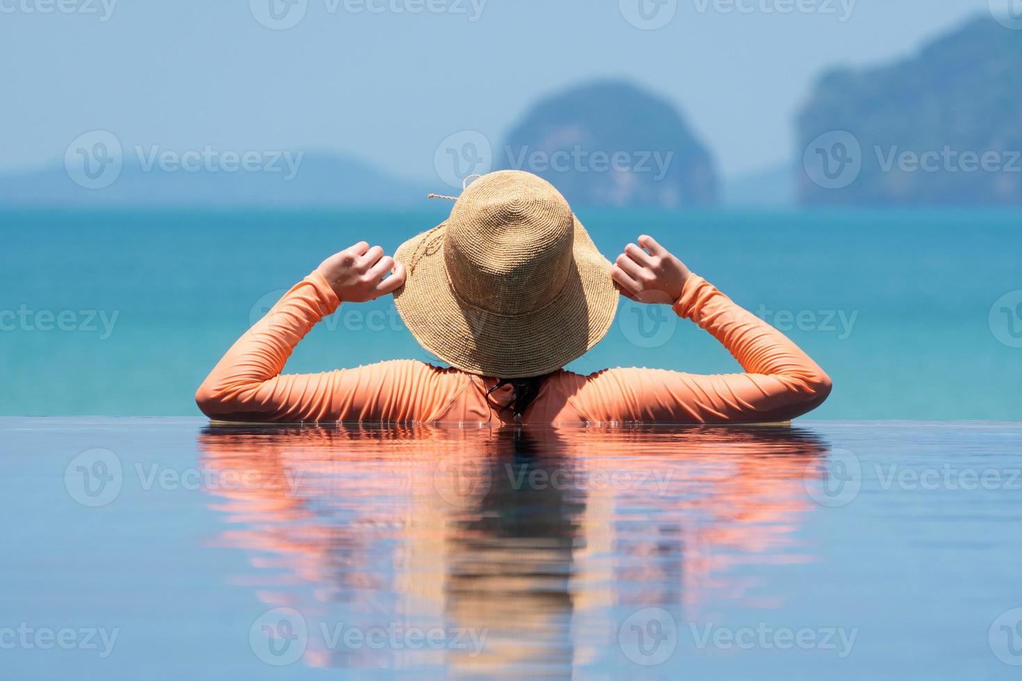 Porträt einer jungen Frau mit Strohhut und Badeanzug, die im blauen Infinity-Pool mit Blick auf den Ozean im Sommerurlaub steht foto