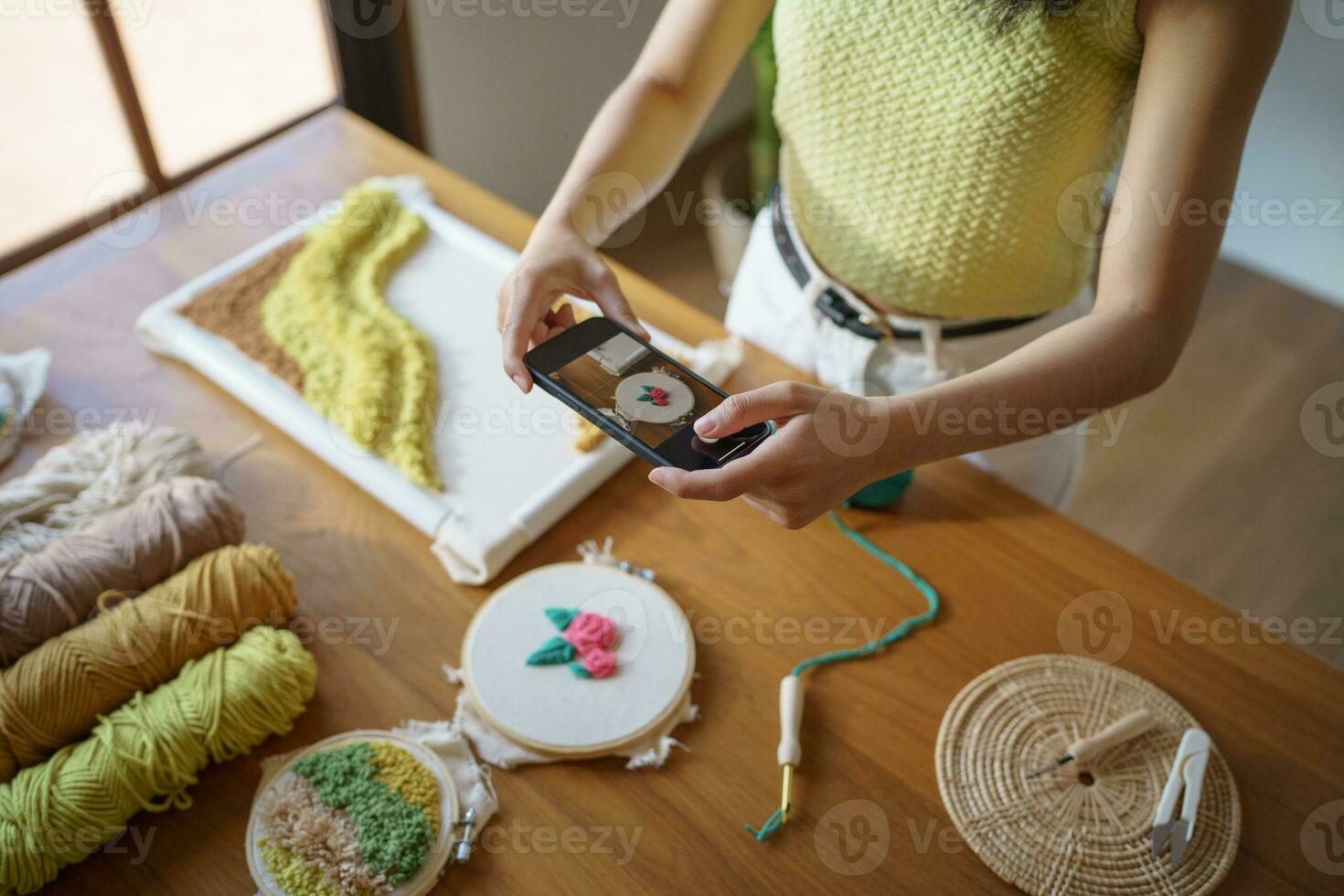 asiatisch Frau nehmen Smartphone Foto schlagen Nadel. Telefon Buchung auf Sozial Netzwerke im Studio Werkstatt. Designer Arbeitsplatz handgemacht Kunst Projekt DIY Stickerei