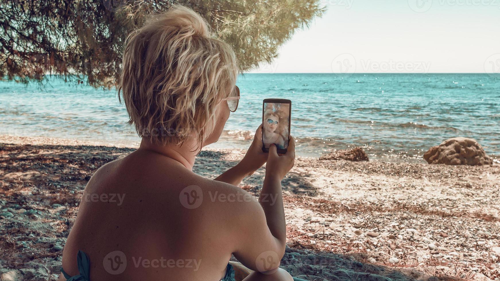 blonde Frau macht das Selfie am Strand. Frau macht das Selfie am wilden Strand im Schatten einer riesigen Kiefer und blickt auf die Ägäis. foto