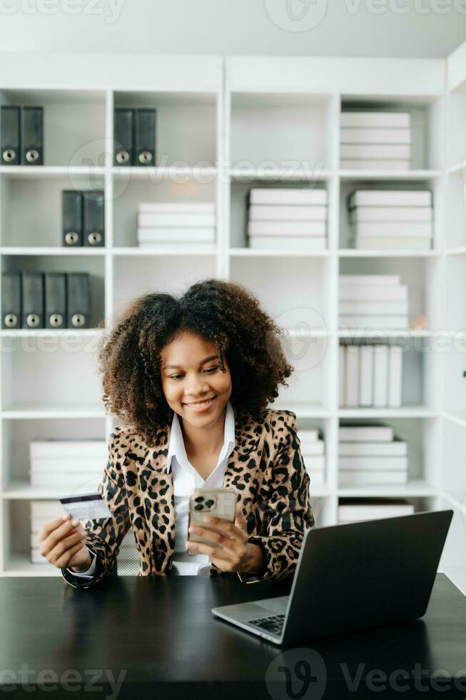 afrikanisch Frau mit Clever Telefon zum Handy, Mobiltelefon Zahlungen online Einkaufen, Omni Kanal,sitzend auf Tisch, virtuell Symbole Grafik Schnittstelle Bildschirm foto