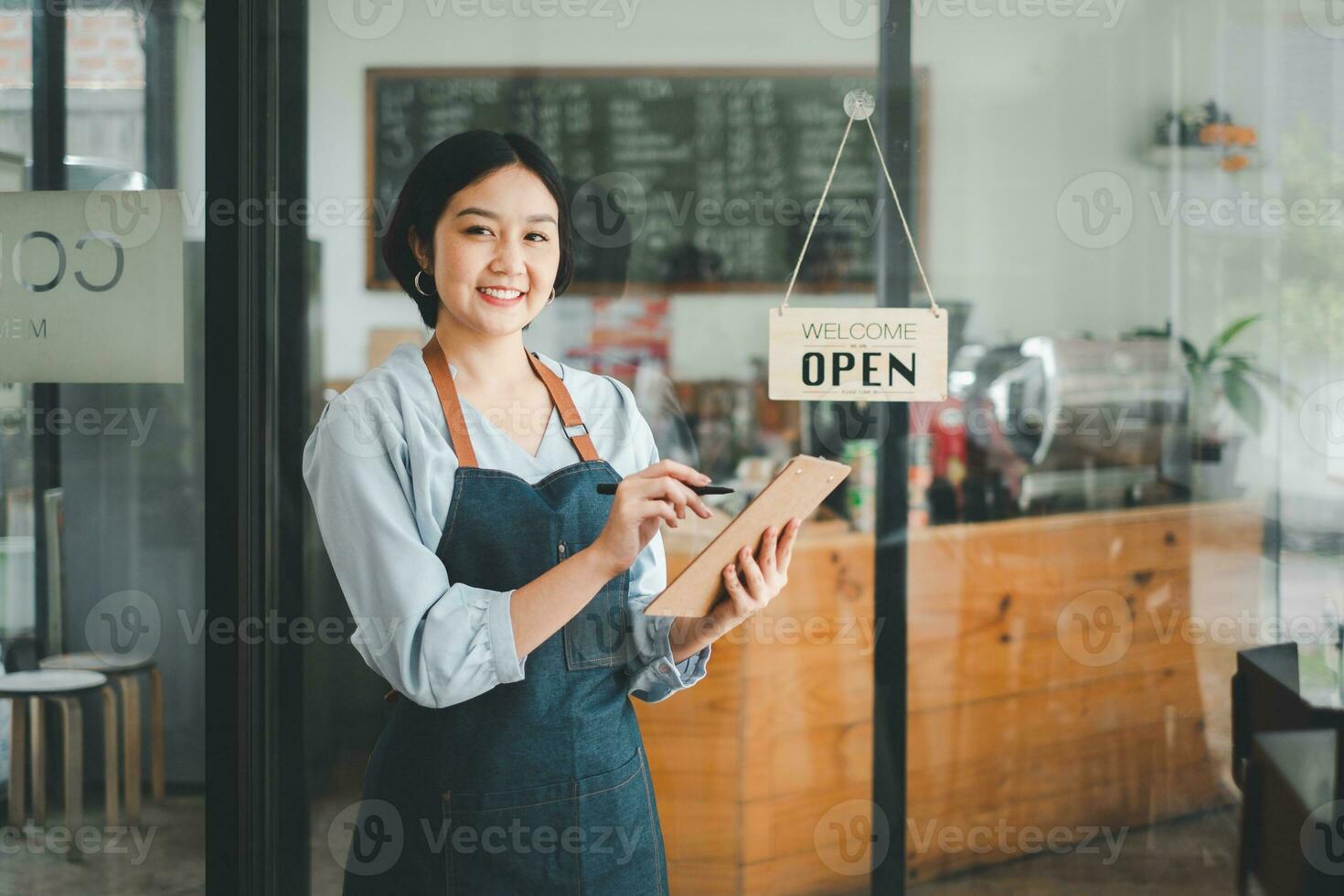 schön jung Barista Frau im Schürze halten bestellen Papier und Stehen im Vorderseite von das Tür von Cafe mit öffnen Zeichen Tafel. Geschäft Inhaber Anfang Konzept. foto