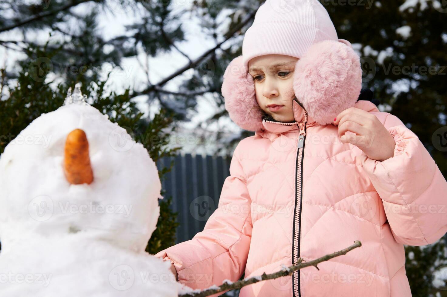 schön süß Kind Mädchen im Rosa Nieder Jacke und flauschige Ohrenschützer, Gebäude Schneemann, genießen glücklich Winter Ferien draußen foto