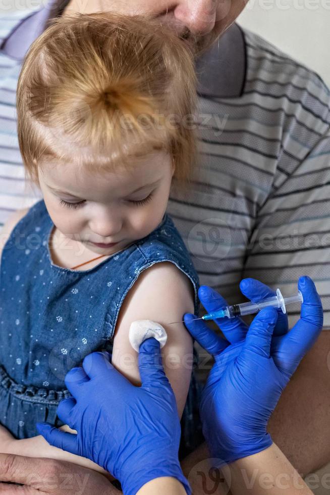 ein kleines mädchen mit ihrem vater in der arztpraxis der klinik wird gegen das coronavirus geimpft. das konzept der impfung, immunisierung, prävention gegen covid-19. foto