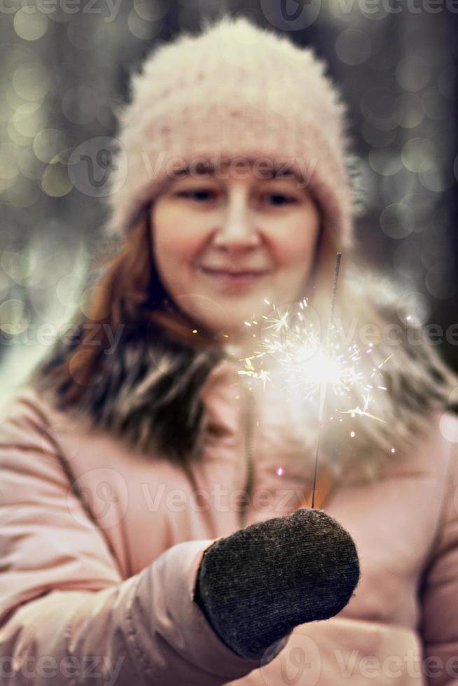 Weihnachtslichter sind helle Spritzer, die in den Händen einer verschwommenen glücklichen Frau im Park brennen. Wunderkerze. Emotionen, Neujahrsstimmung.Winterferien foto