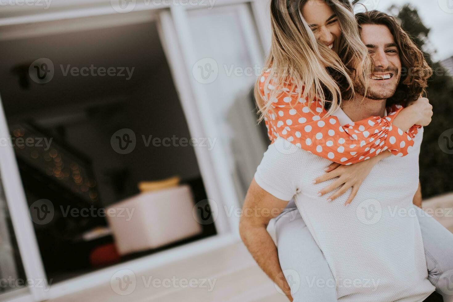 gut aussehend lange Haar Mann Tragen das jung Frau auf seine zurück im Vorderseite von Backstein Haus foto