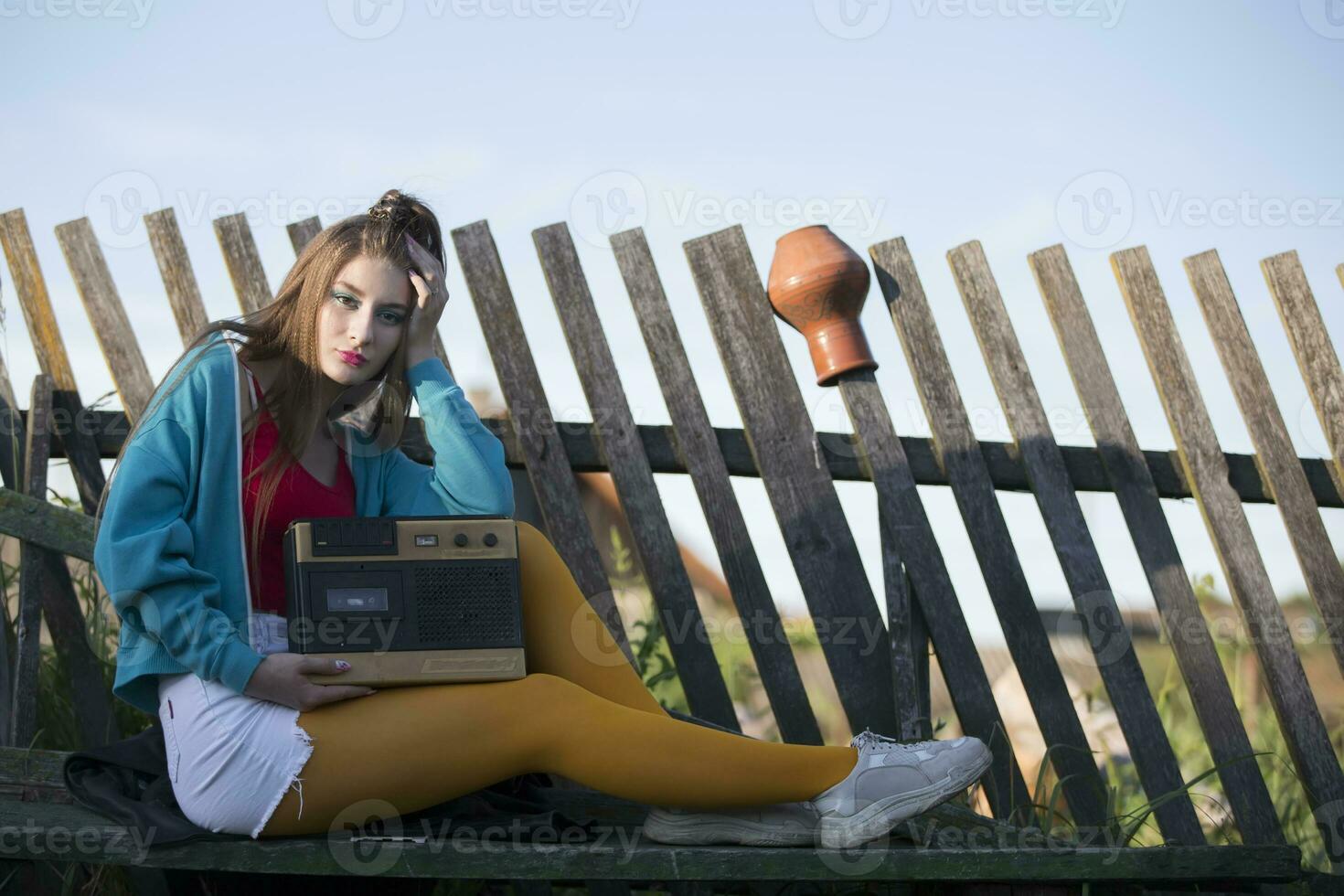 ein schön Land Mädchen im hell Kleider sitzt auf ein hölzern Bank mit ein Kassette Recorder. Frau im das Stil von das 90er. foto