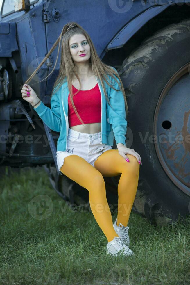 ein schön Land Mädchen im das Stil von das 90er Jahre im hell Kleider steht in der Nähe von ein Traktor. foto