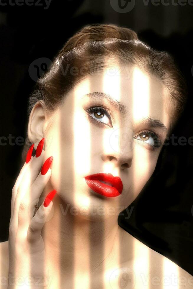 ein Modell- mit rot Lippenstift und Nagel Polieren ist beleuchtet durch das hell Licht von das Jalousie auf ein dunkel Hintergrund. das Gesicht von ein schön gepflegt Mädchen mit sauber Haut. foto