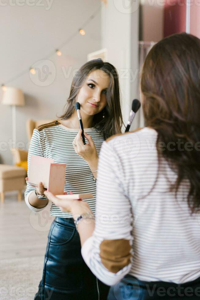 schön Mädchen setzt erröten auf ihr Gesicht mit ein bilden Bürste während Stehen beim Zuhause im ein Zimmer und suchen im das Spiegel. kaukasisch Mädchen im ein gestreift Hemd tut ihr besitzen bilden foto