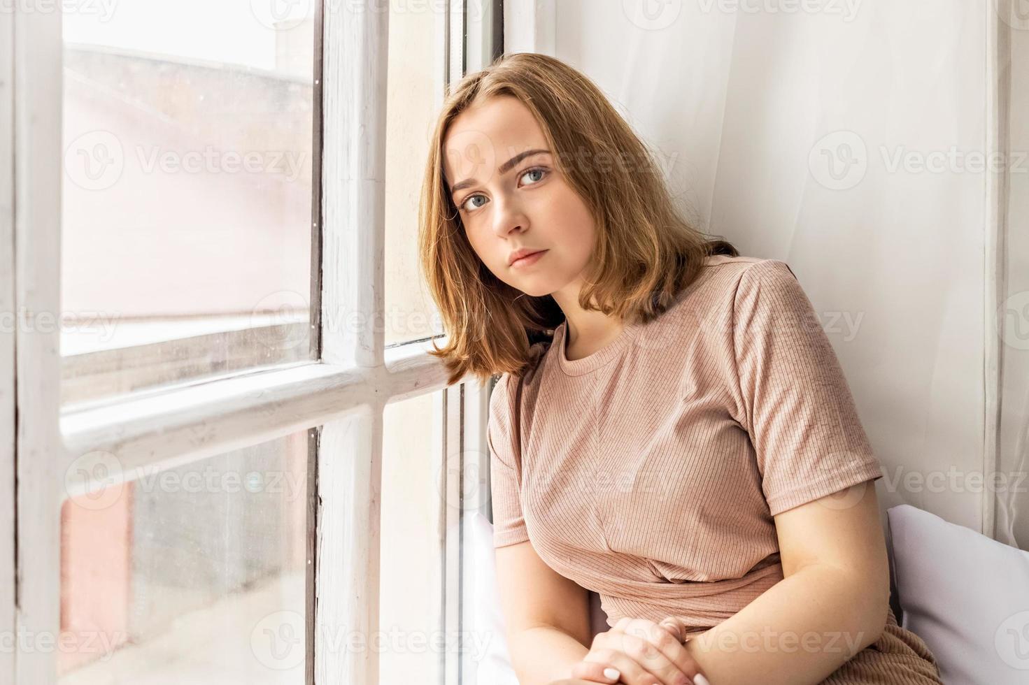 Nachdenkliches schönes junges Mädchen sitzt auf einem Fensterbrett in der Nähe des Fensters. Porträt eines Teenagers allein zu Hause. Menschen, Lifestyle, Freizeit und Entspannungskonzept. foto