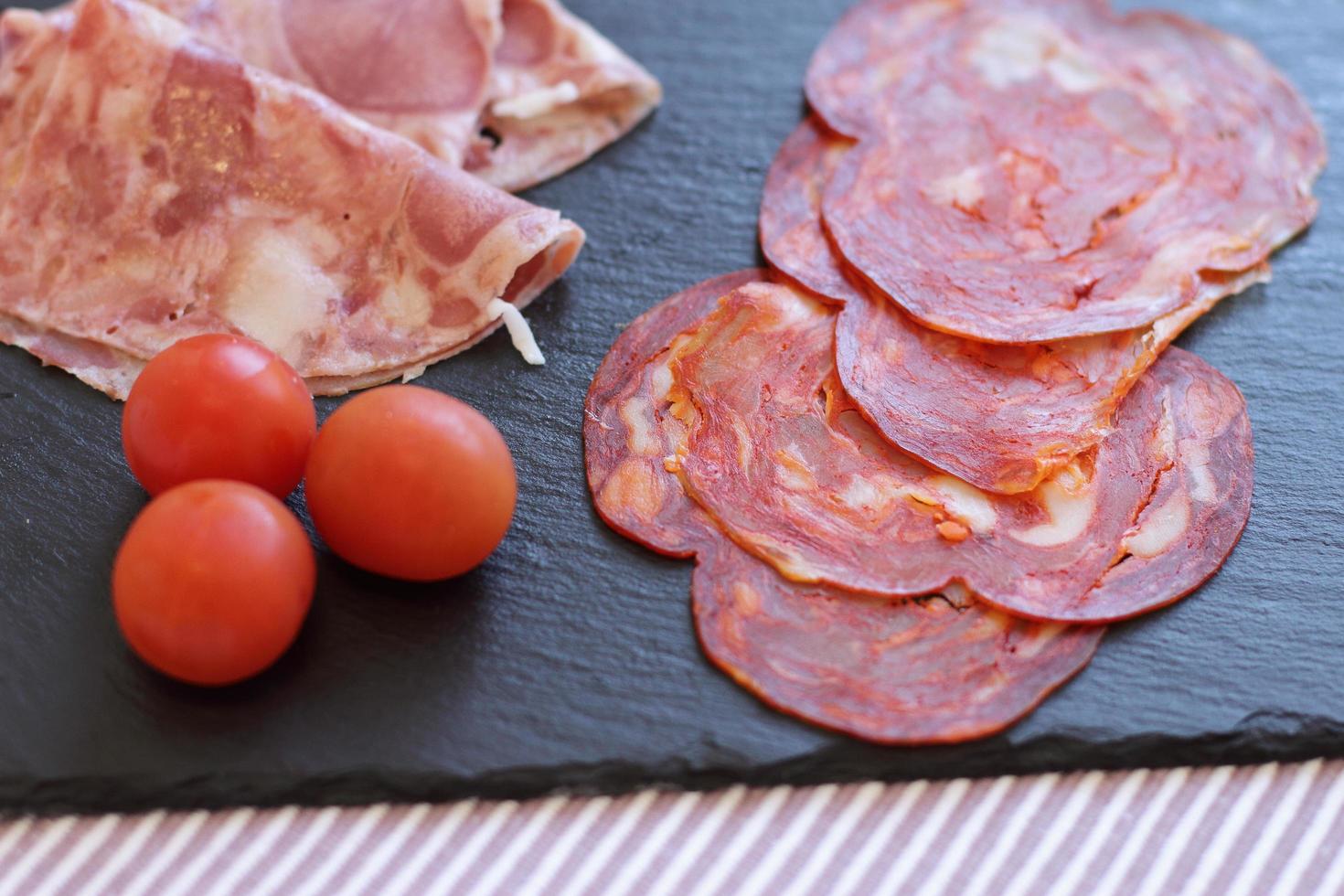 frische Scheiben Salami und Chorizo auf schwarzem Brett mit drei Tomaten Kirsche foto