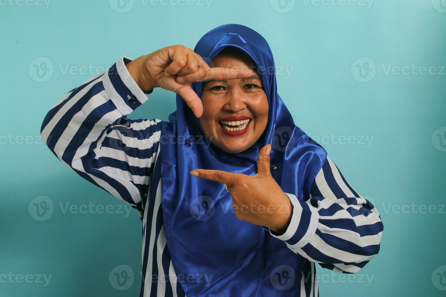ein inspiriert mittleren Alters asiatisch Frau, im ein Blau Hijab und gestreift Shirt, macht ein Rahmen Geste, suchen das perfekt Winkel oder Inspiration zu Erfassung ein Moment im ein Foto. isoliert auf ein Blau Hintergrund foto