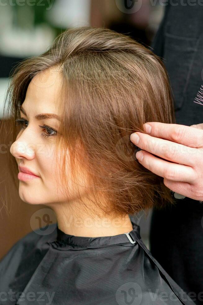 männlich Friseur funktioniert auf das Frisur von das jung kaukasisch Brünette Frau beim ein Haar Salon. foto