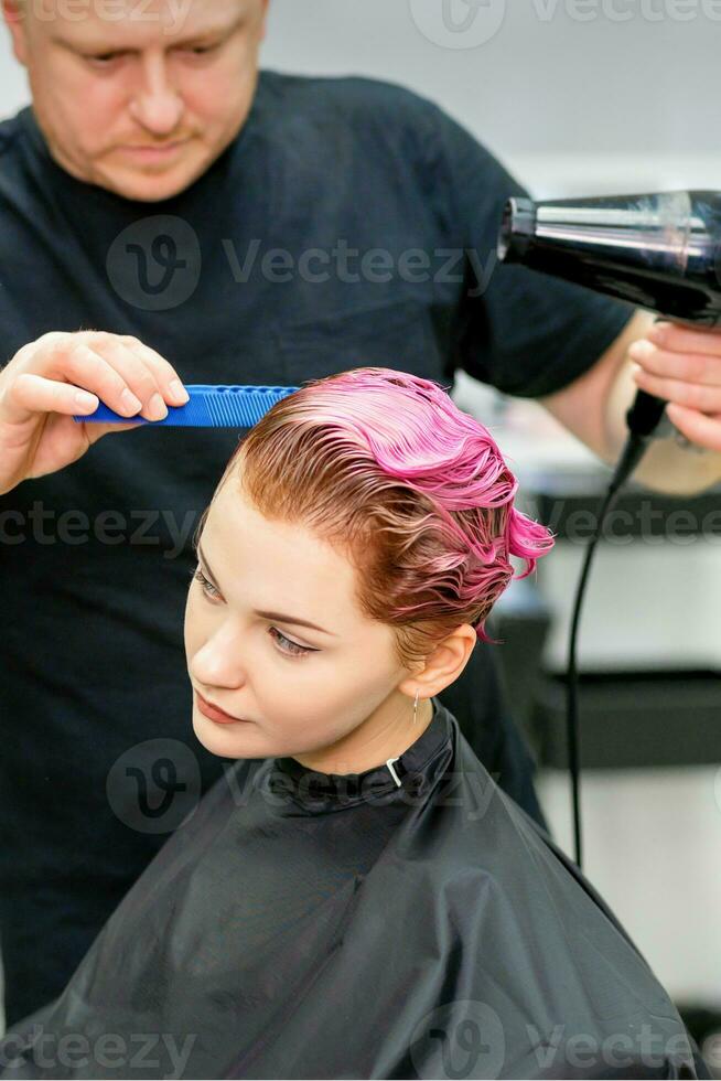 ein Friseur ist Trocknen das Rosa Haar von das jung Frau im ein Schönheit Salon. foto