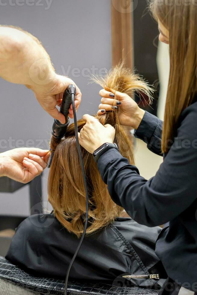 das Rückseite Aussicht von zwei Friseure sind Eisstockschießen Haar zum ein jung Frau mit elektrisch Haar Eisen im ein Schönheit Salon. foto