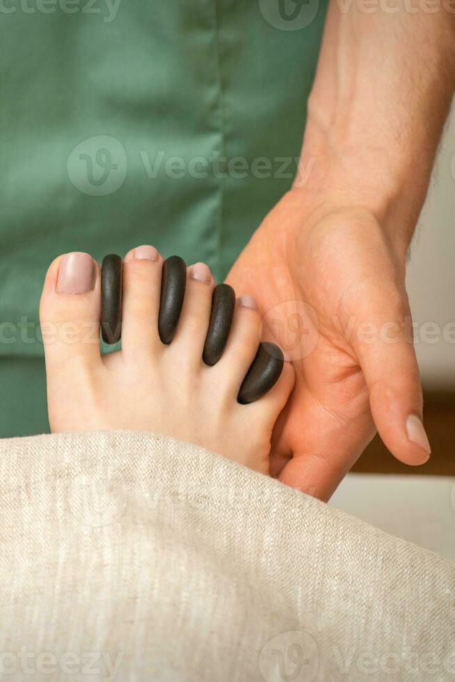 Füße Massage mit schwarz Kieselsteine zwischen das weiblich Zehen im Hände von das Masseur im das Spa. foto