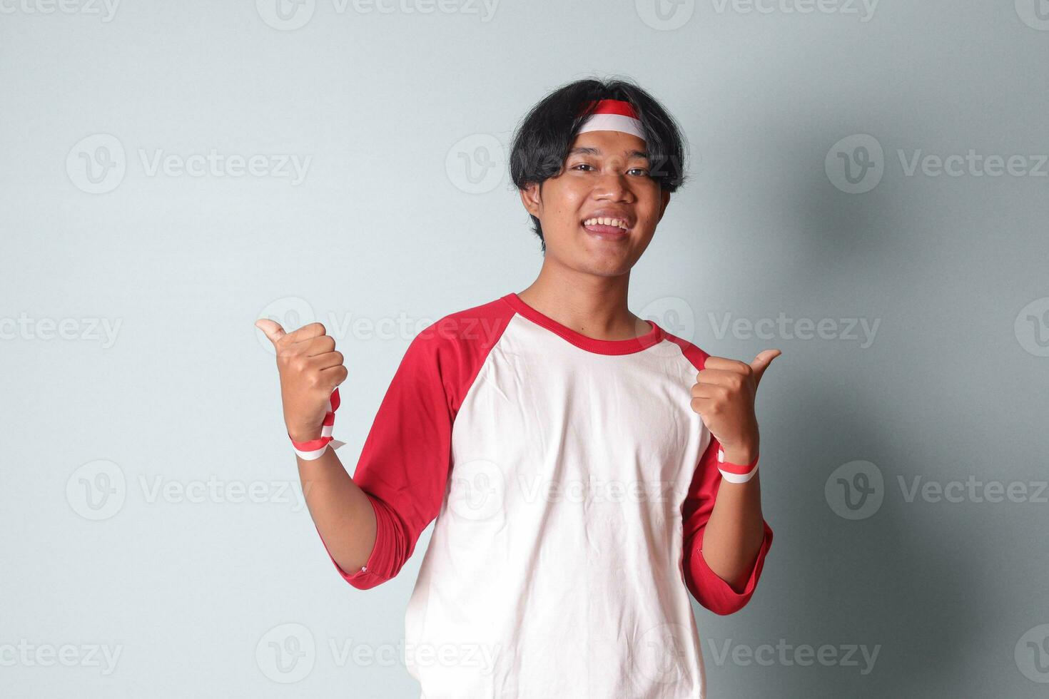 Porträt von attraktiv asiatisch Mann im T-Shirt mit rot und Weiß Band auf Kopf, zeigen gut Job Hand Geste mit Daumen hoch. isoliert Bild auf grau Hintergrund foto