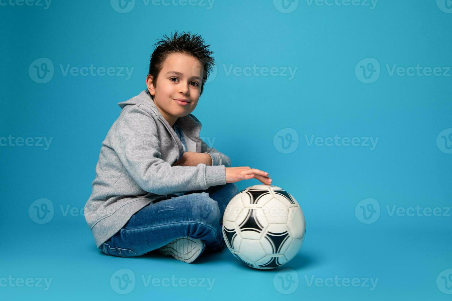 gut aussehend Kind Sitzung in der Nähe von ein Fußball Ball auf Blau Oberfläche foto