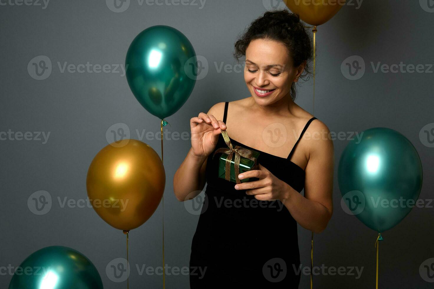 attraktiv heiter Frau im Abend Balck Kleid hält ein Geschenk im Hände und lächelt posieren gegen aufgeblasen bunt Grün metallisch und golden Luft Luftballons auf grau Hintergrund mit Kopieren Raum zum Anzeige foto