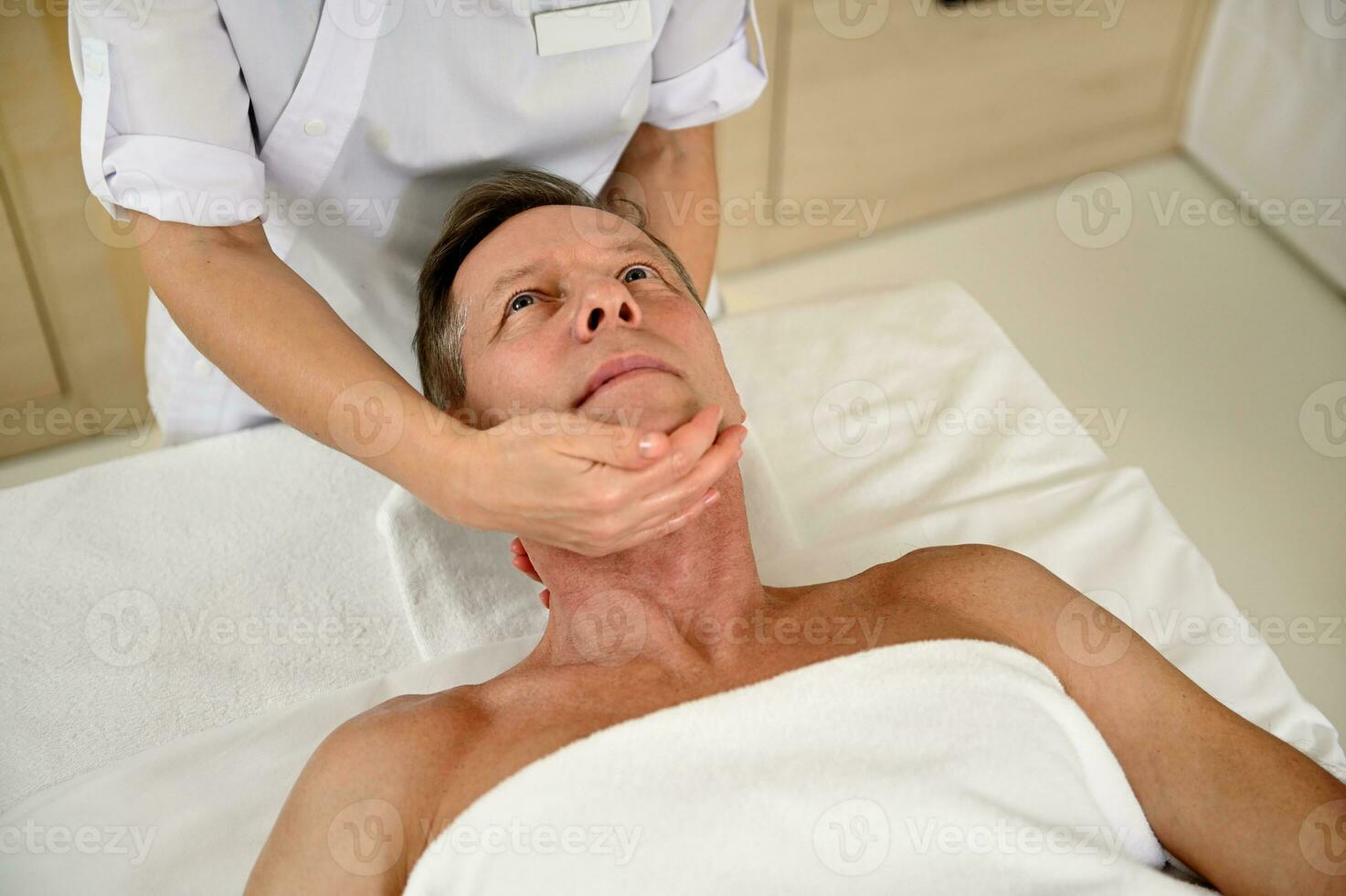 hoch Winkel Aussicht von gut aussehend reifen Mann Empfang therapeutisch Massage beim ein Wellness Klinik. Nahansicht von Physiotherapeut massieren Hals von Mitte alt Klient beim ayurvedisch Spa Center. Alternative Therapie foto