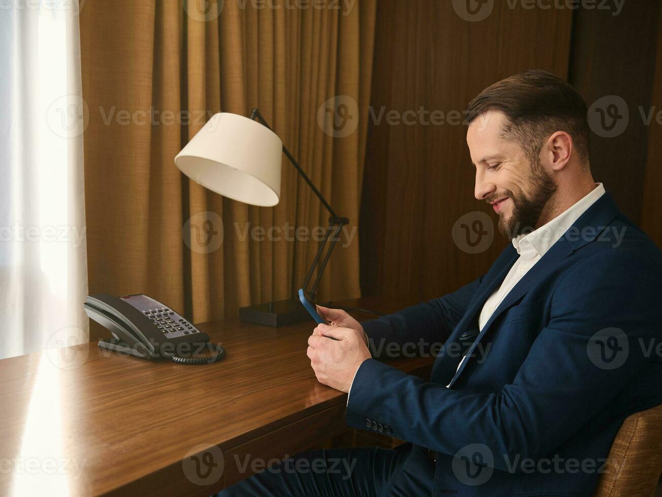 Seite Porträt von ein gut aussehend Geschäft Reisender, Geschäftsmann, Unternehmer, Investor im dunkel passen ruhen im Hotel Zimmer, lächelt während chatten auf Handy, Mobiltelefon Telefon während Geschäft Ausflug foto