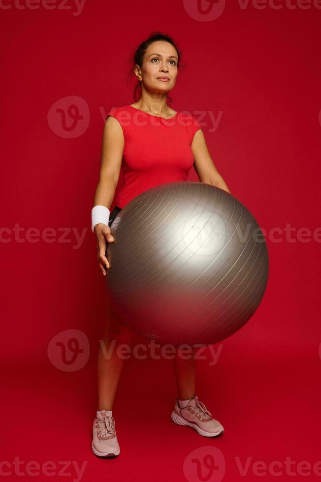 attraktiv muskulös mittleren Alters spanisch Frau im Sportbekleidung halten passen Ball, isoliert Über rot Hintergrund mit Kopieren Raum zum Werbung Text foto