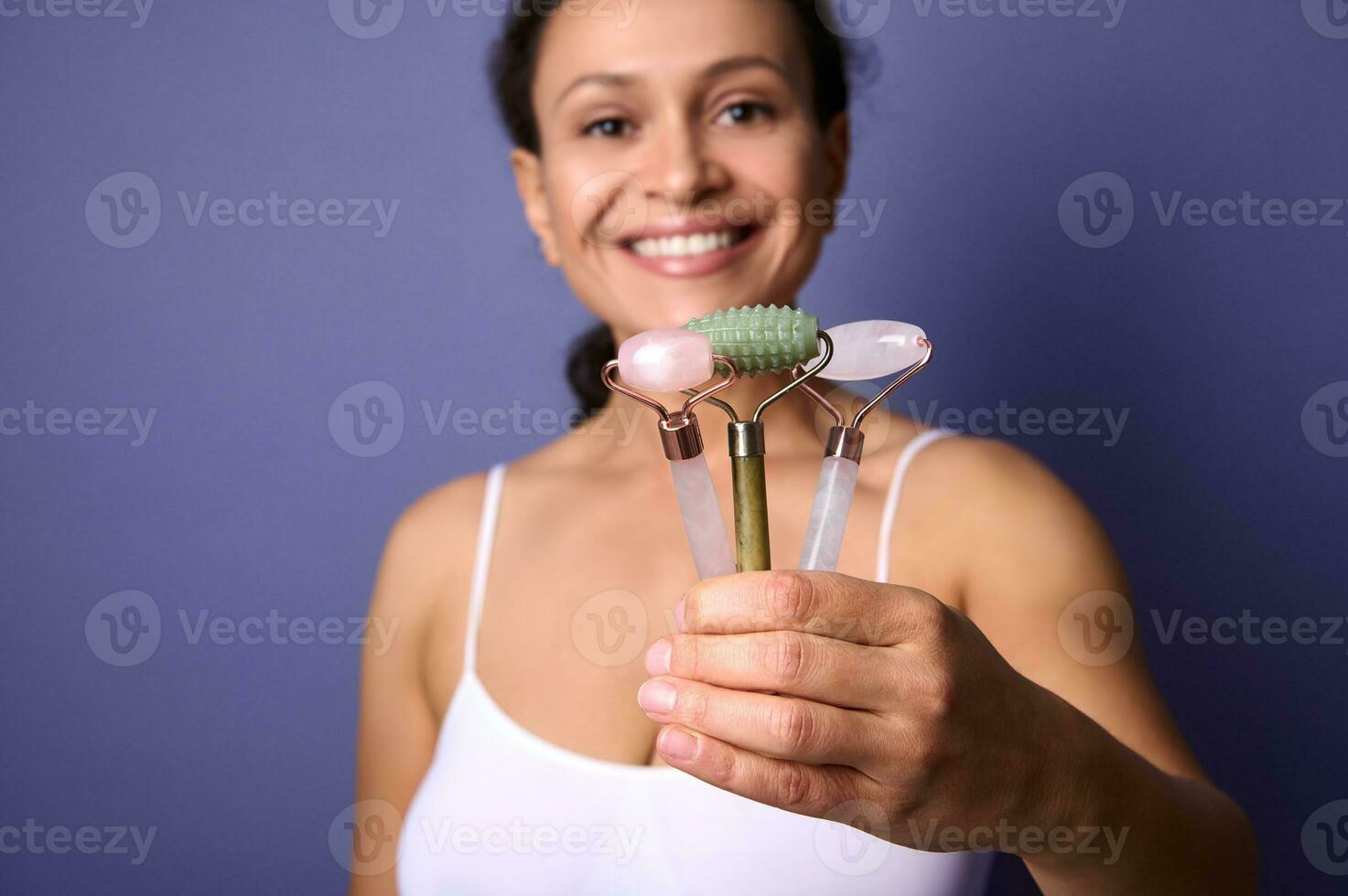 Fokus auf bunt ayurvedisch Jade Stein Walze Massagegeräte im das Hand von heiter lächelnd Mitte alt ziemlich Frau posieren gegen violett Hintergrund. Gesicht und Haut Pflege Konzept mit Kopieren Raum. Nahansicht foto