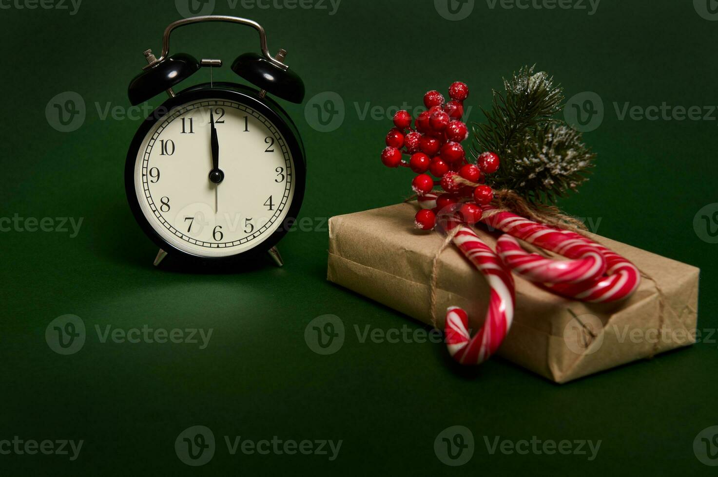 Nahansicht von schwarz Alarm Uhr mit Mitternacht auf das wählen, Süss gestreift Lutscher, Süßigkeiten Stöcke und Stechpalme auf Weihnachten Geschenk im Kunst Verpackung Papier isoliert auf Grün Hintergrund mit Kopieren Raum zum Anzeige foto