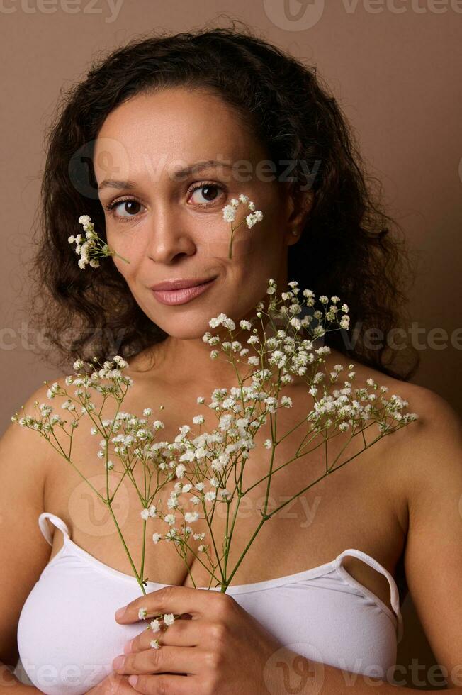 Mode Schönheit Porträt von attraktiv sinnlich feminin Frau mit Gypsophila Weiß Zweig selbstbewusst suchen beim Kamera, posieren gegen Beige farbig Hintergrund. Körper, Haut Pflege, Damen Tag Konzept foto