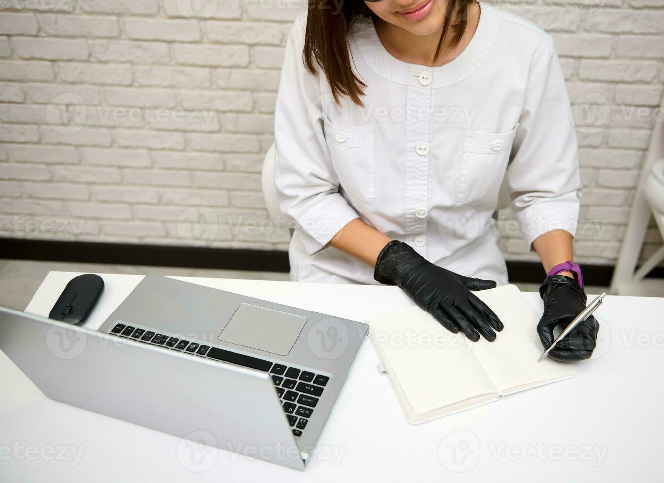 hoch Winkel Aussicht von ein Frau Arzt, Allgemeines Praktiker im Weiß medizinisch Uniform Sitzung beim Schreibtisch im Krankenhaus Schreiben und Beratung geduldig online auf Laptop, lächelt selbstbewusst suchen beim Kamera foto