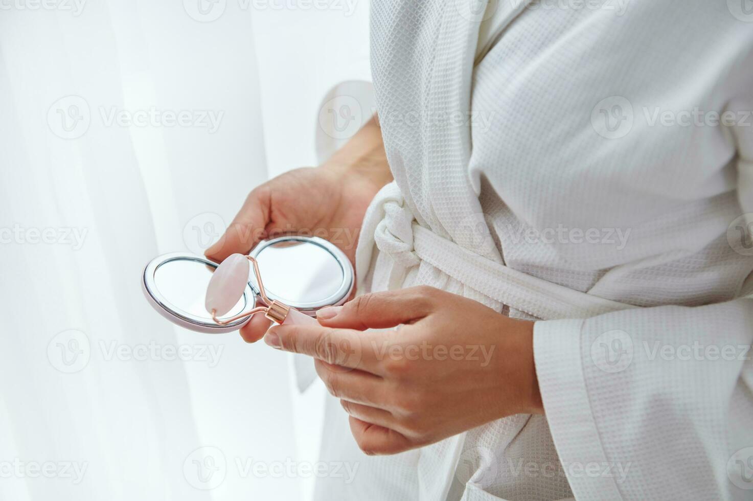 Nahansicht von weiblich Hände halten klein kosmetisch Spiegel und Jade Stein Walze Massagegerät. abgeschnitten Bild von ein Frau im Weiß Bademantel mit Jade Walze ein ein Spiegel im ihr Hände foto
