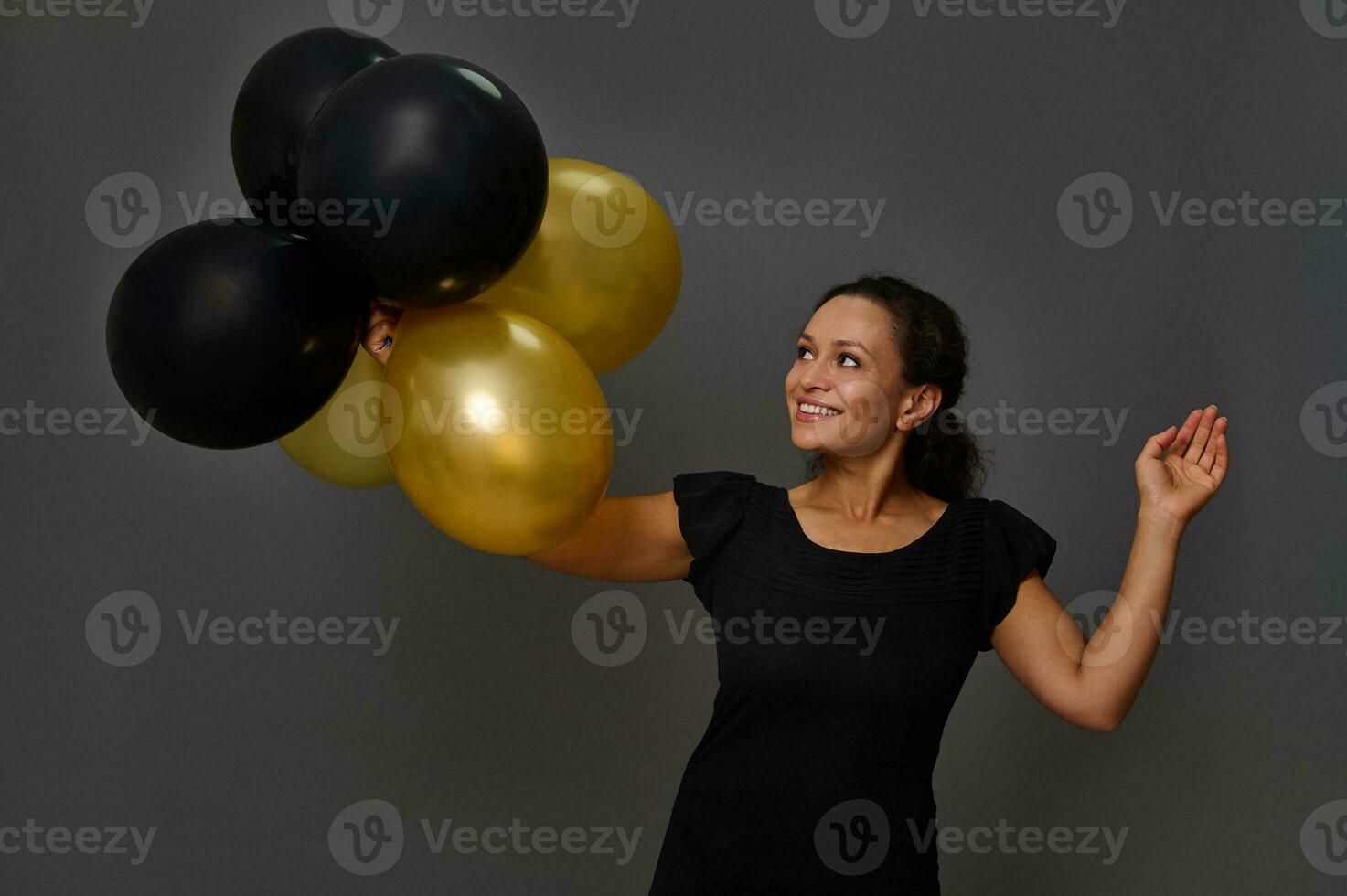 heiter ziemlich spanisch Frau mit schön Lächeln posiert mit aufgeblasen schwarz und Gold Luft Luftballons gegen grau Mauer Hintergrund mit Kopieren Raum. Konzept von schwarz Freitag und Gold Anerkennung Einkaufen Karte foto