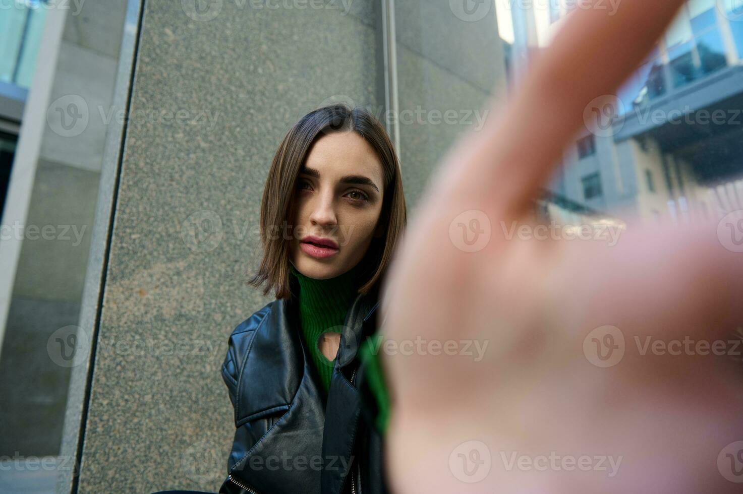 Fokus auf ein schön attraktiv Brünette Frau halten ausgestreckt Hand auf unsichtbar Smartphone zum Herstellung Selbstporträt, und suchen beim Kamera, Kopfschuss, Nahansicht foto