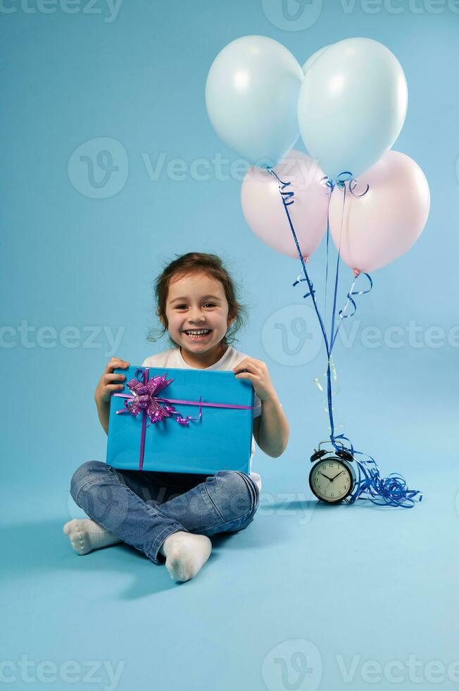froh Mädchen Kind süß lächelnd beim das Kamera Sitzung auf ein Blau Hintergrund Nächster zu ein Alarm Uhr und Luftballons und halten ein Blau Geschenk mit ein Rosa Bogen foto