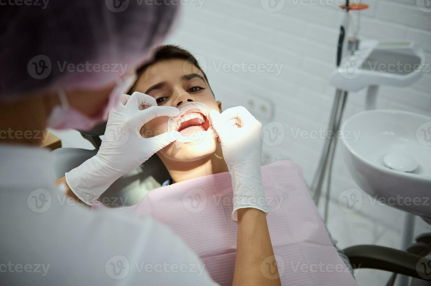 Overhead Aussicht von pädiatrisch Zahnarzt Putten Wange Dental Retraktor auf Kind geduldig Vor Oral Behandlung. Schule Junge beim das Zahnarzt Termin. Dental Karies Behandlung und gesund Zähne Konzept. foto