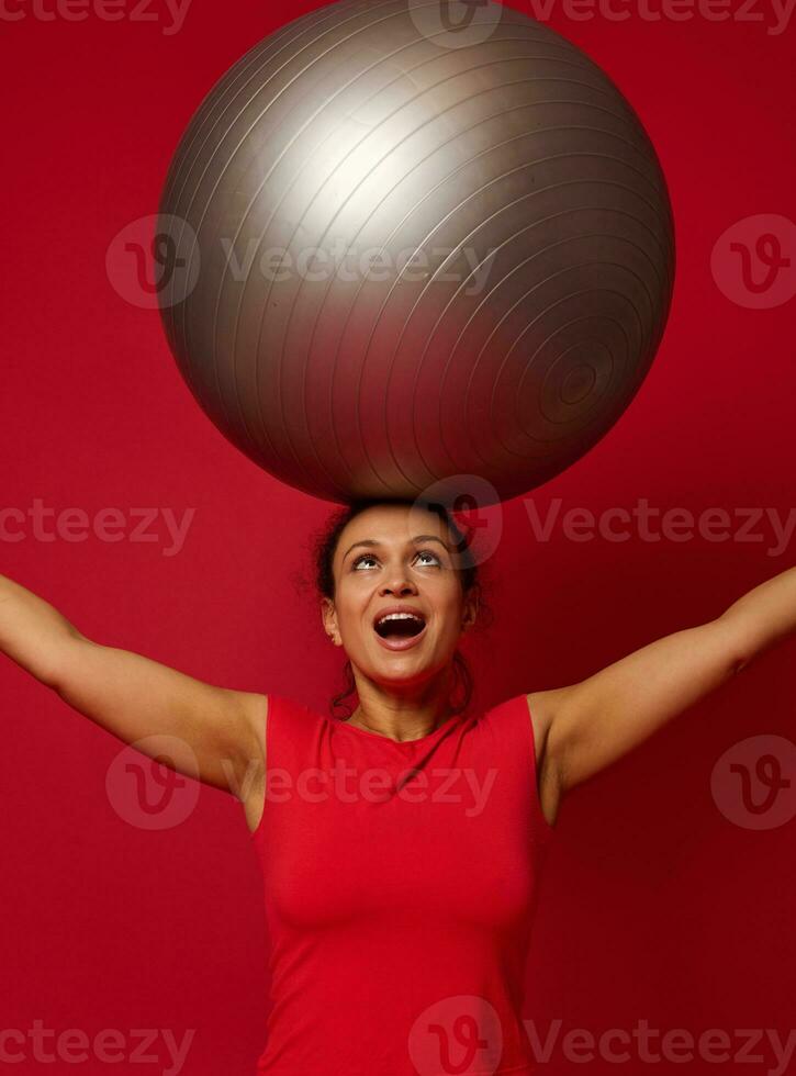 Porträt von ein Fitness glücklich afrikanisch amerikanisch passen Frau Stehen halten ein passen Ball im ihr angehoben Waffen über ihr Kopf. Kopieren kostenlos Raum zum Text. foto