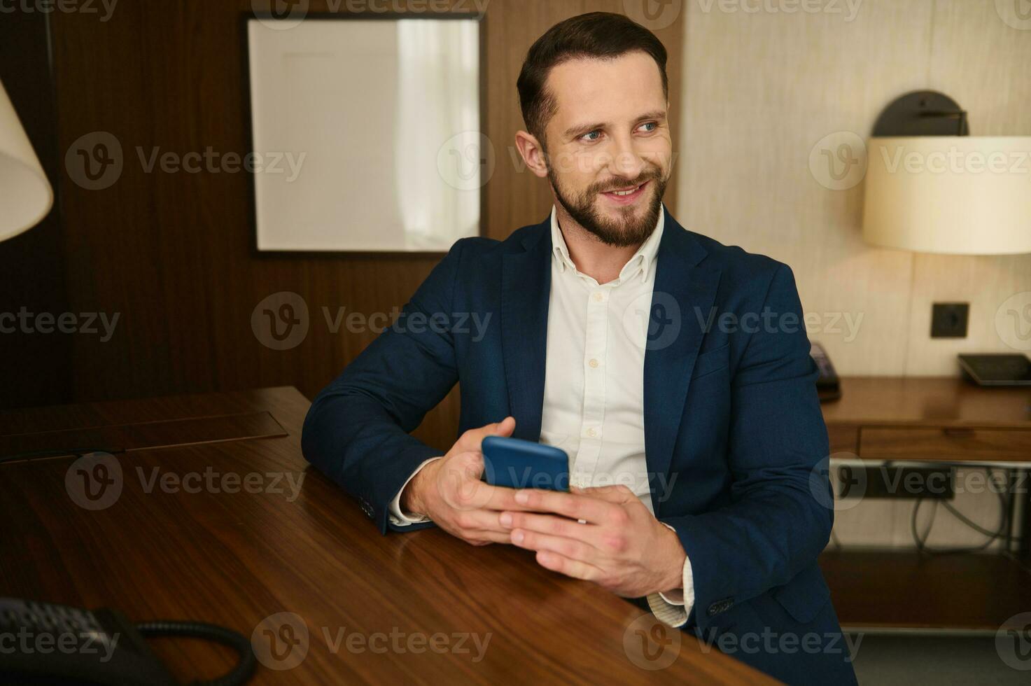 lächelnd attraktiv Geschäftsmann mit Handy, Mobiltelefon Telefon suchen beiseite Sitzung beim ein Tabelle während ruhen im Hotel Zimmer während Geschäft Ausflug foto