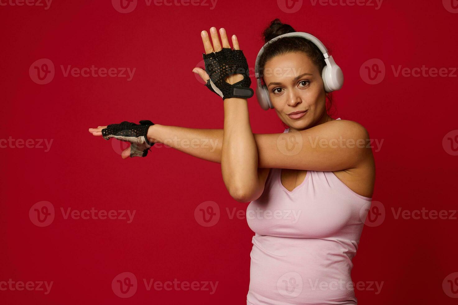 zuversichtlich aktiv mittleren Alters aktiv Frau im Sportbekleidung und Kopfhörer Dehnen Waffen, suchen beim Kamera gegen rot Hintergrund mit Kopieren Anzeige Raum foto