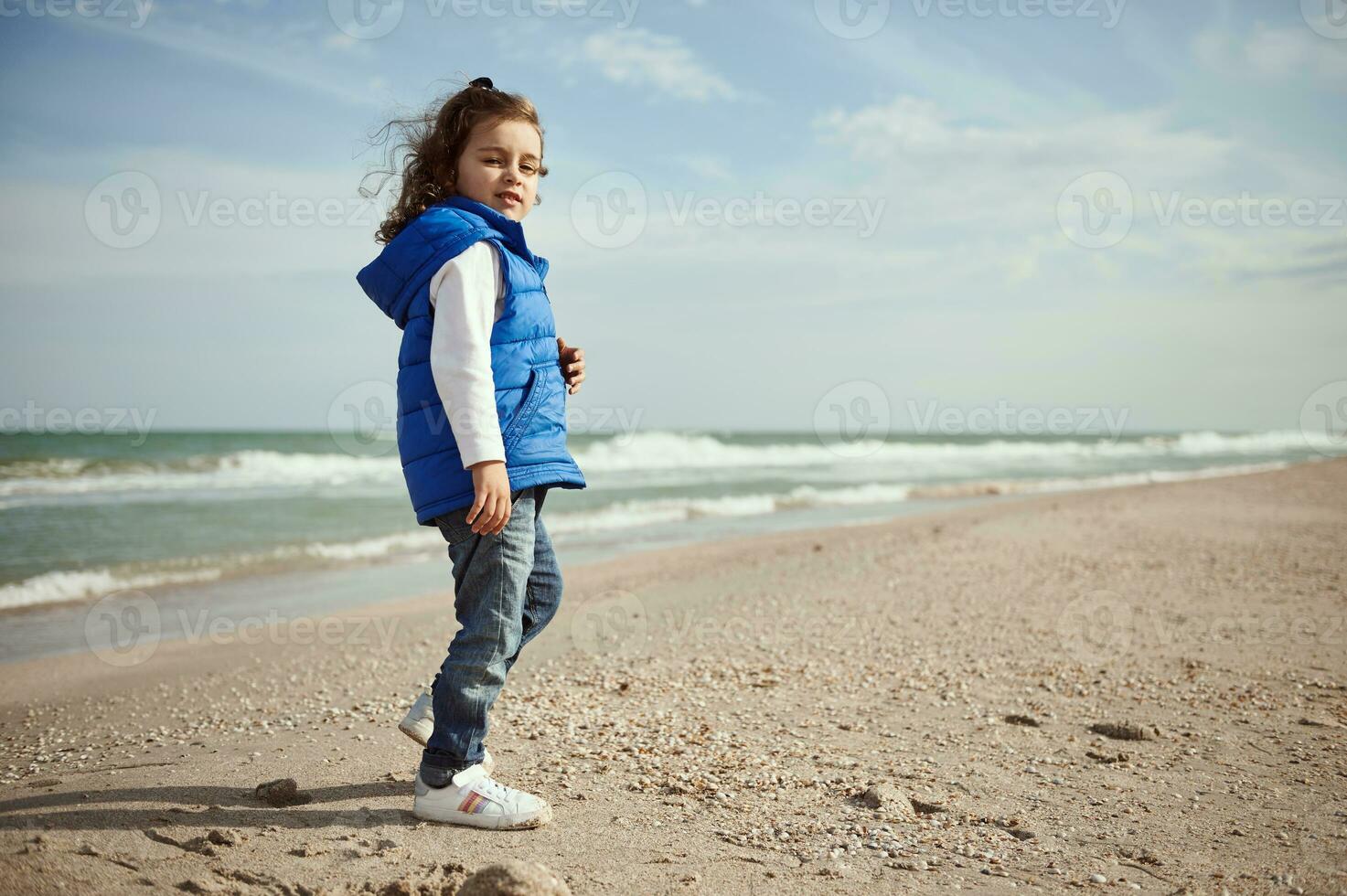 schön wenig Kind suchen beim Kamera durch Schulter während Stehen auf das Strand. International Kinder- Tag. glücklich Kindheit Konzept. foto
