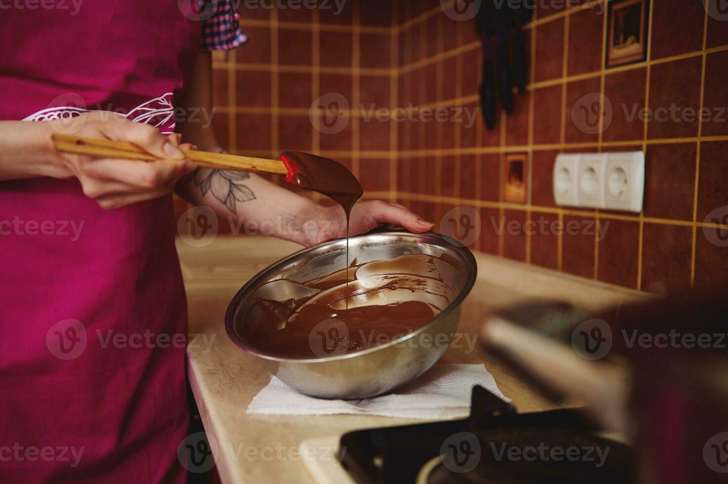 Nahansicht von ein Konditor im Rosa Schürze Mischen geschmolzen Schokolade Masse im Schüssel mit ein hölzern Spatel foto