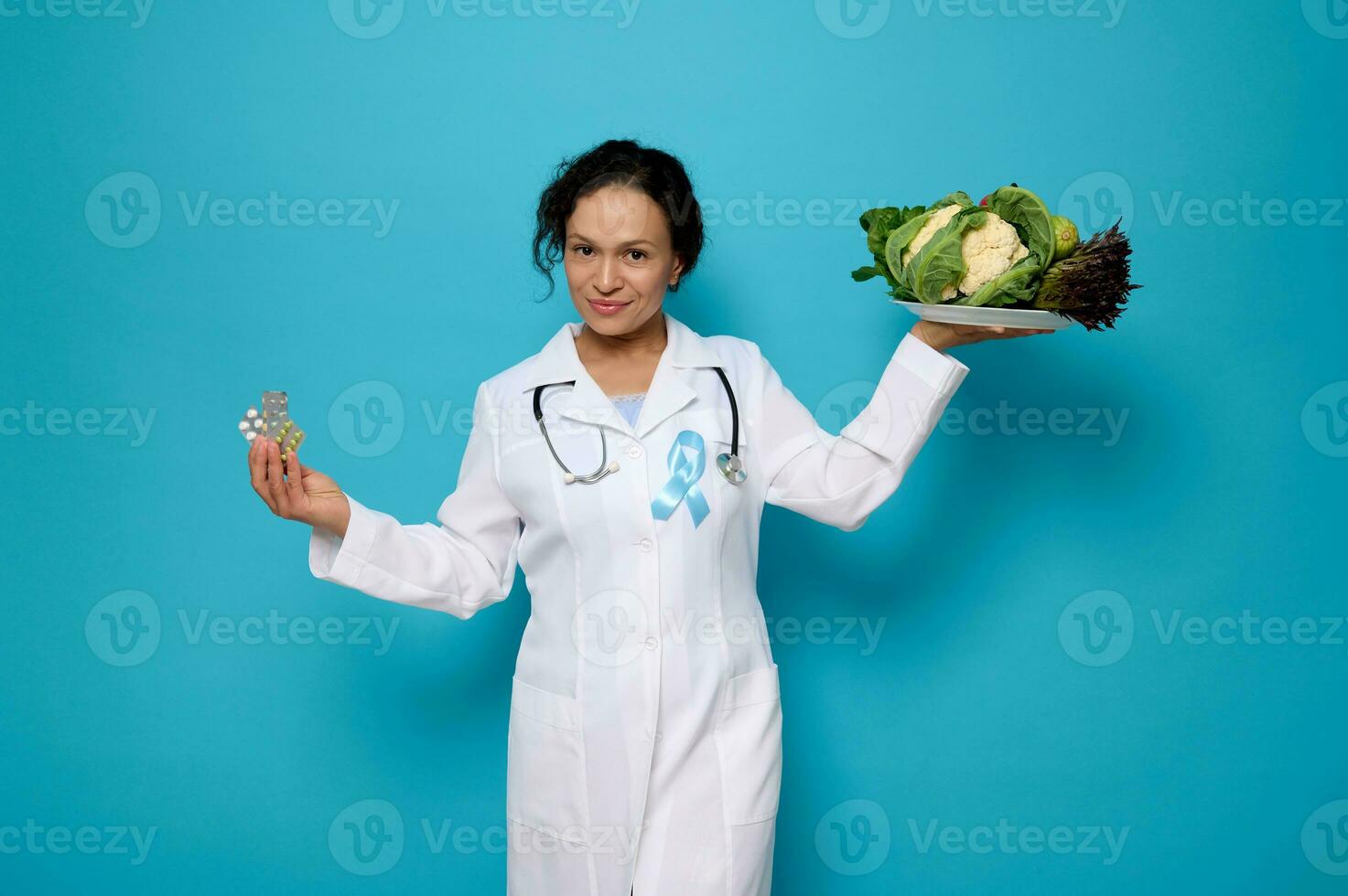 schön zuversichtlich weiblich Arzt im Weiß medizinisch Kleid mit Blau Band, Symbol von Welt Diabetes Bewusstsein Tag, hält ein Teller mit gesund vegan Essen und Blase mit Medikamente im Hände foto