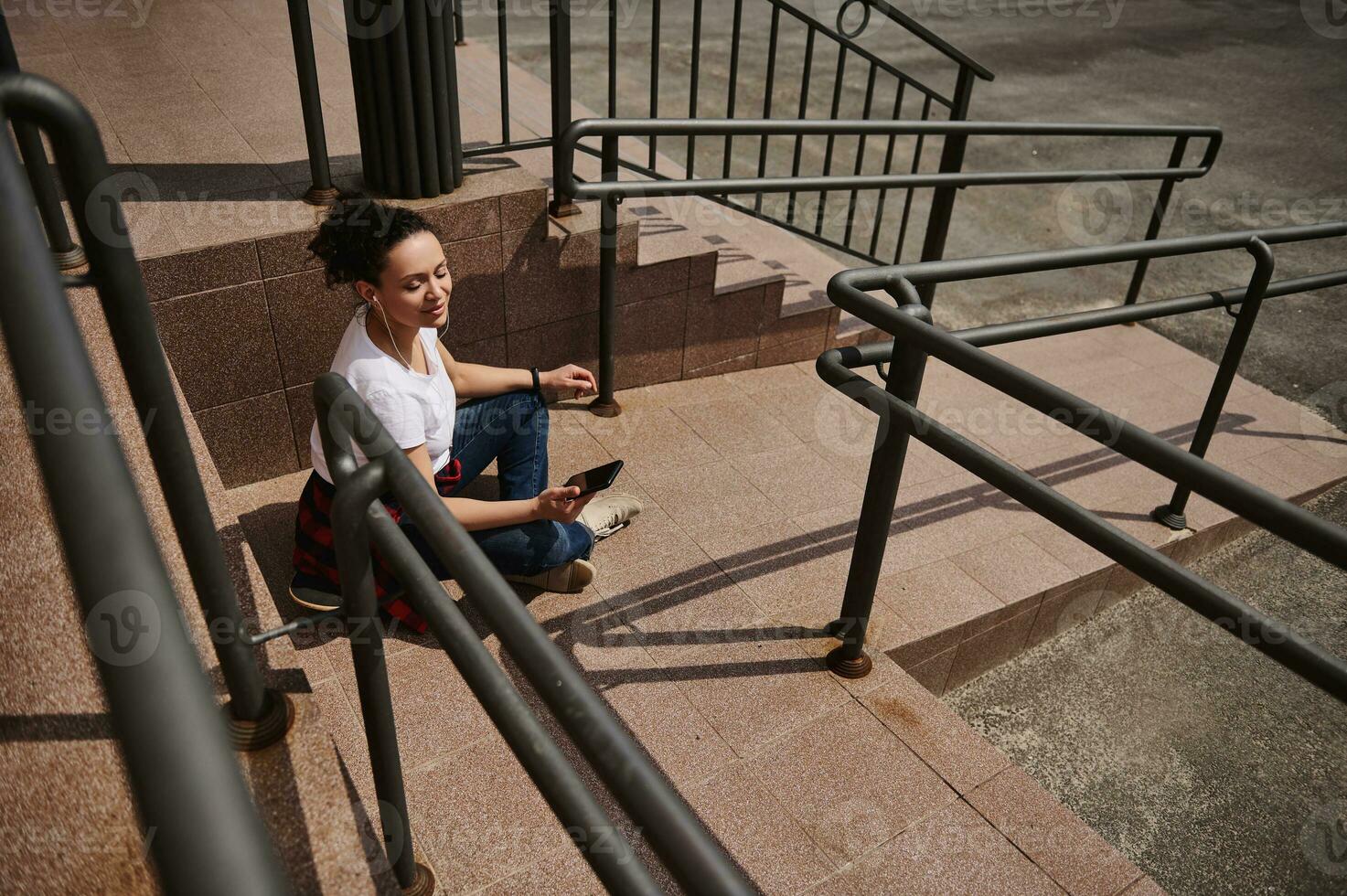 jung Latein amerikanisch Frau im Kopfhörer mit Handy, Mobiltelefon Telefon während Sitzung auf Skateboard auf Hintergrund von Schritte foto