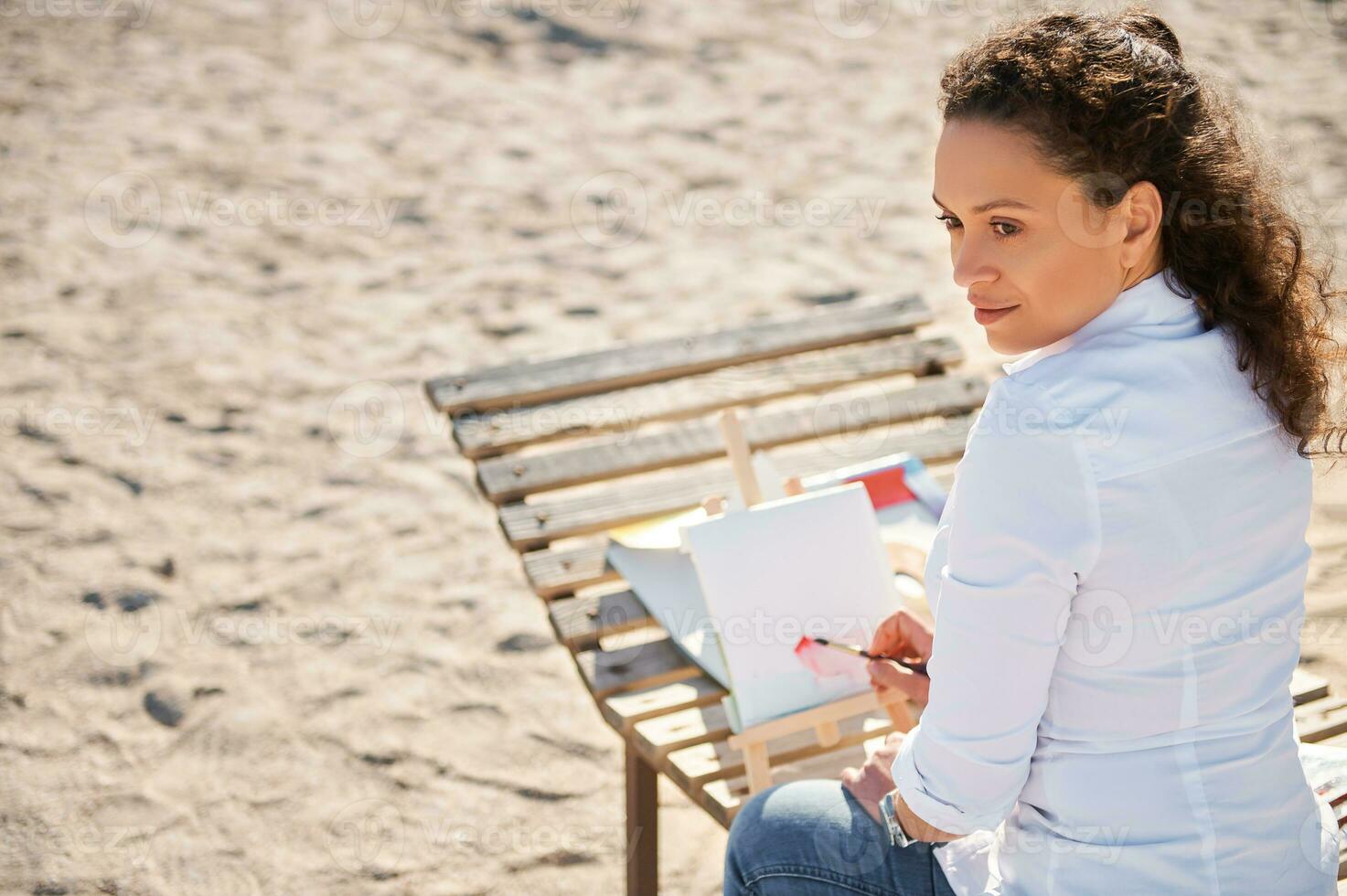 schön Frau suchen in das Entfernung und Zeichnung mit Aquarelle Sitzung auf ein hölzern Chaise Salon auf das Sitzung auf einsam Strand foto