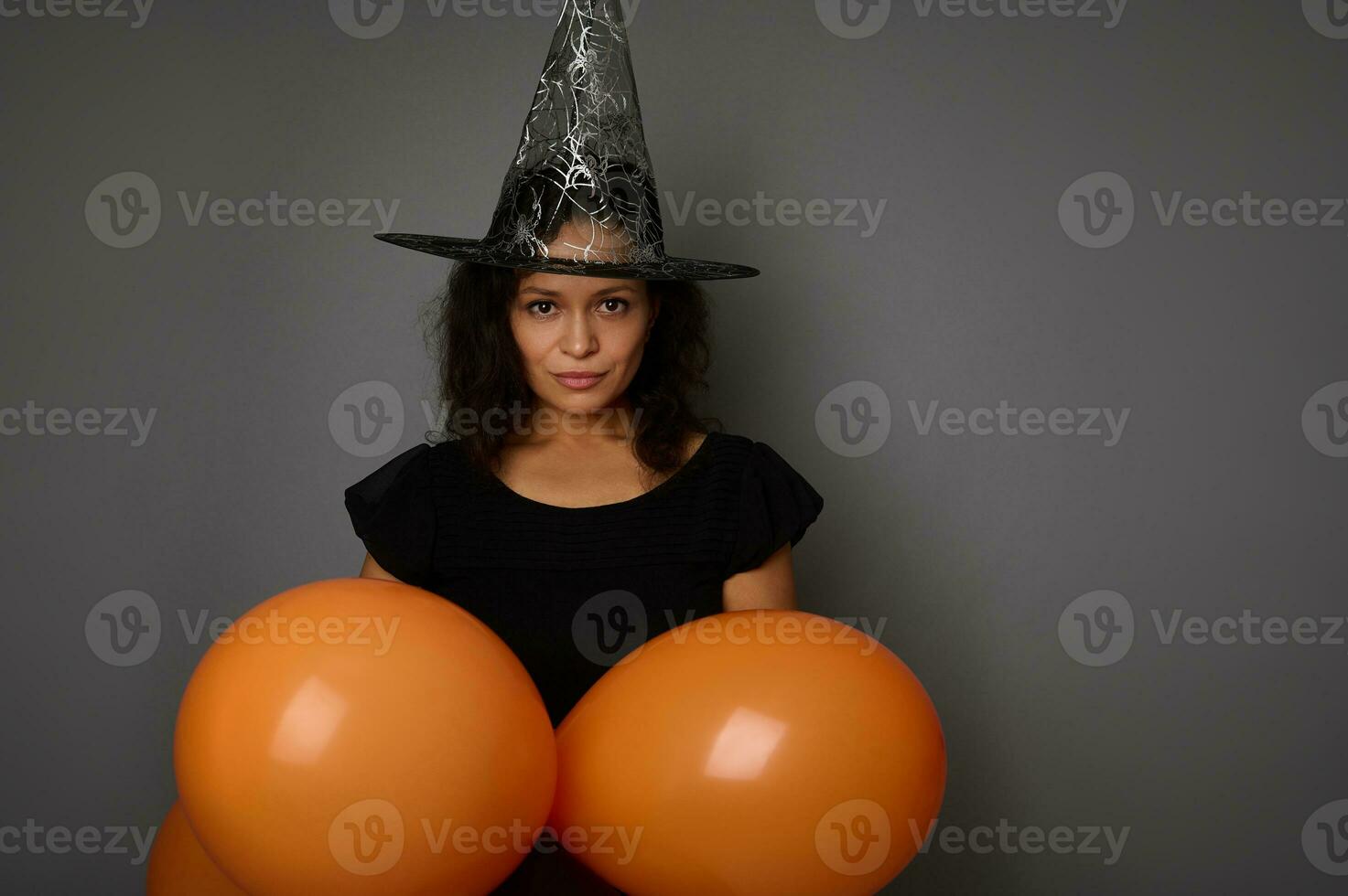 charmant mysteriös gemischt Rennen Frau gekleidet im schwarz Hexe Karneval Halloween Kostüm und Magier Hut, hält Orange bunt Luft Luftballons, sieht aus beim Kamera, isoliert Über grau Hintergrund, Kopieren Raum. foto