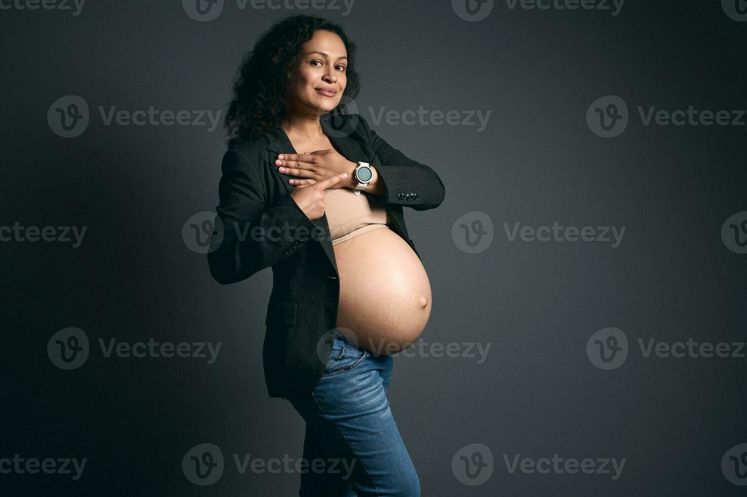 stilvoll ethnisch schwanger Frau im grau Blazer und beiläufig Jeans, zeigt an ihr Clever Uhr lächelt beim Kamera, Studio Hintergrund foto